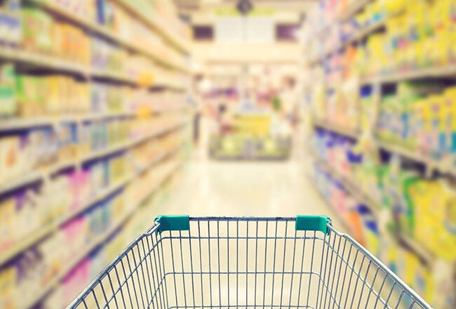 A shopping cart is pushed down an aisle in a supermarket, which is filled with colorful products on shelves, slightly blurred in the background.