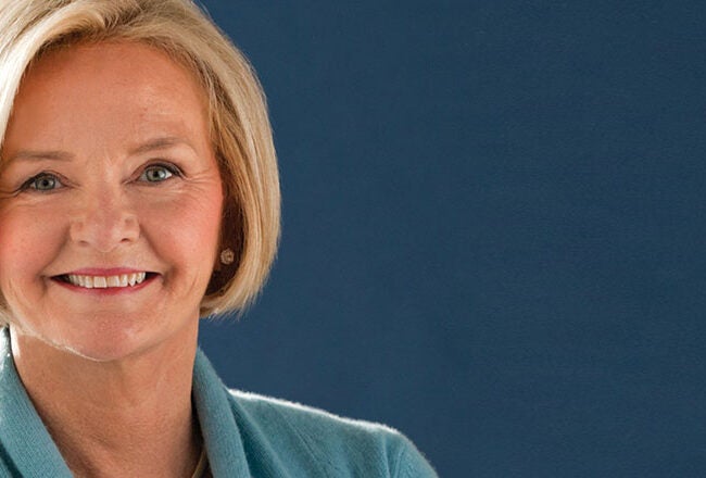 A person with short, light hair and a warm smile, wearing a light blue sweater, poses against a solid dark blue background.