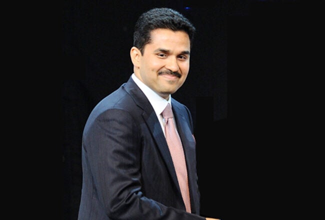 A headshot of a person wearing a dark suit with a white shirt and pink tie, set against a black background, smiling and looking forward.