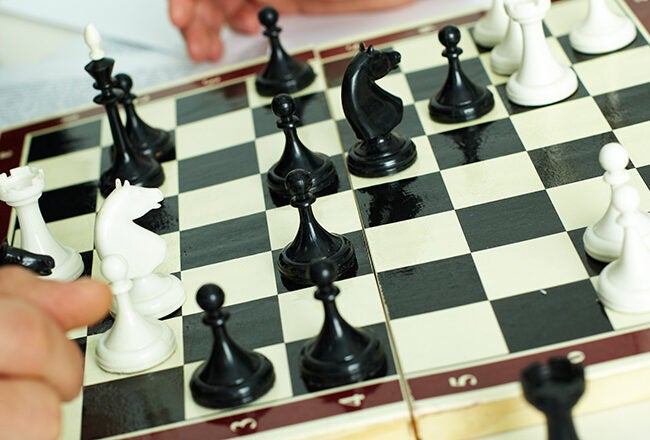 A close-up of a chess game in progress on a board, with both black and white pieces visible. A person''s hand is moving a piece.