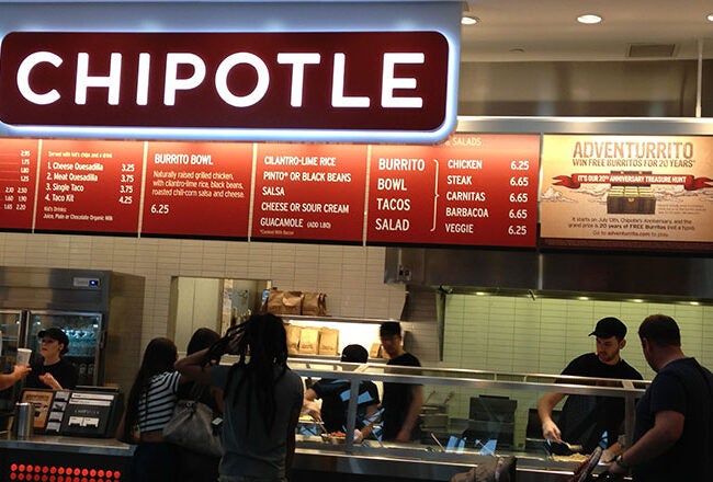 People ordering food at a Chipotle Mexican Grill counter with menu boards displaying various options like burritos and tacos.