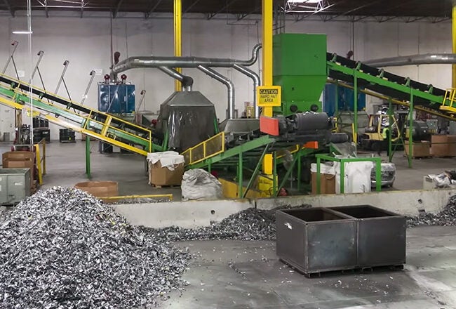 An indoor recycling facility with conveyor belts, machinery, and piles of sorted materials, including metals. The setting appears industrial.