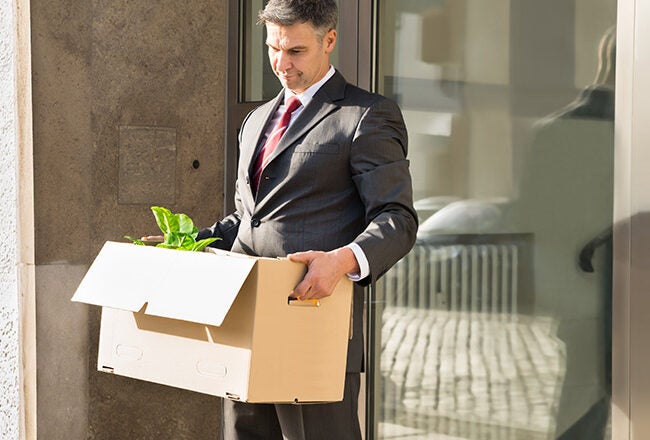 Person in a suit carrying a cardboard box with office supplies and a potted plant, possibly indicating someone leaving a job or being laid off.