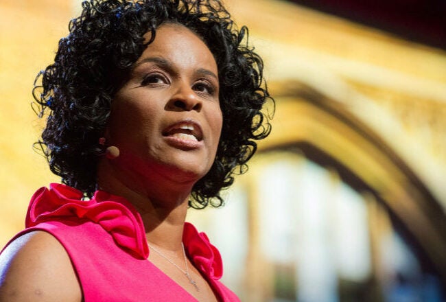 A person with curly hair is speaking on stage, wearing a pink top. They have a microphone headset. The background features a blurred architectural design.