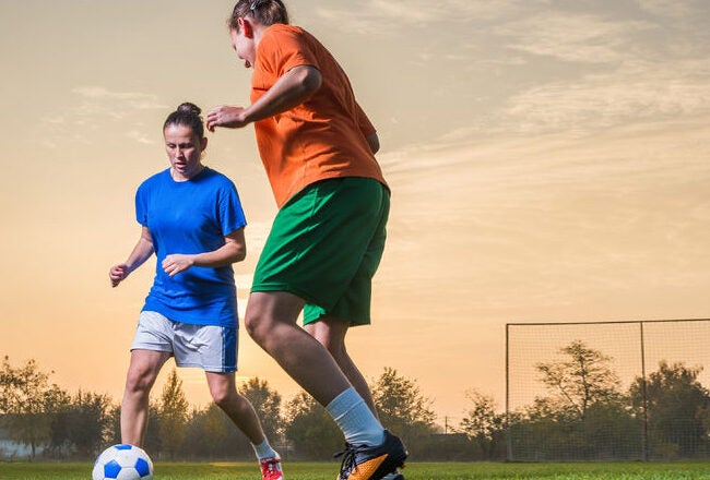 Two people playing soccer on a grass field at sunset, one in a blue shirt and the other in an orange shirt, focusing on the ball.