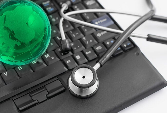 A stethoscope on a laptop keyboard with a green globe hologram, representing global digital healthcare or telemedicine.