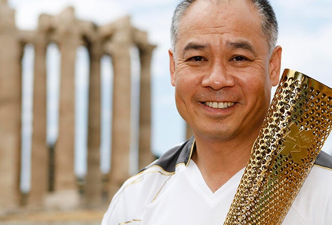 A person holding an Olympic torch stands in front of ancient Greek columns.