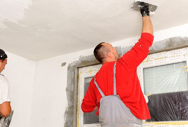 Two people plastering a ceiling in a room, both wearing overalls. A window is covered with protective plastic.