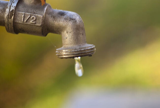 A close-up of a metal water tap with a single droplet forming at the spout, set against a blurred outdoor background, symbolizing water conservation.