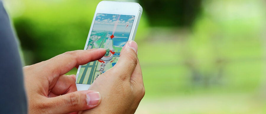 Person playing a game on a smartphone outdoors, with a blurred green background. The game screen shows a virtual map with various icons.