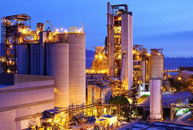 An industrial facility at night, illuminated with lights, featuring large silos and towers. The background includes a body of water and distant mountains.