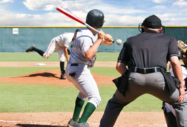 Baseball game scene showing a batter ready to hit a pitch, with a catcher and umpire behind them. The pitcher is in motion on the mound.