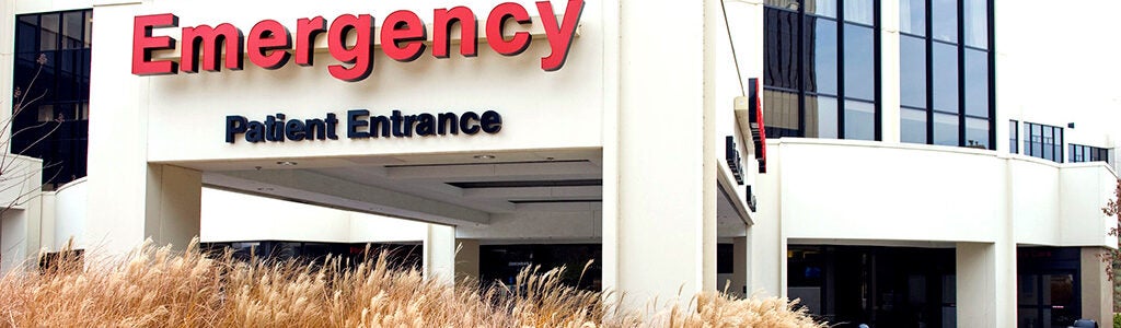 Entrance to an emergency room with a sign reading "Emergency Patient Entrance" on a hospital building. Tall grasses in the foreground.