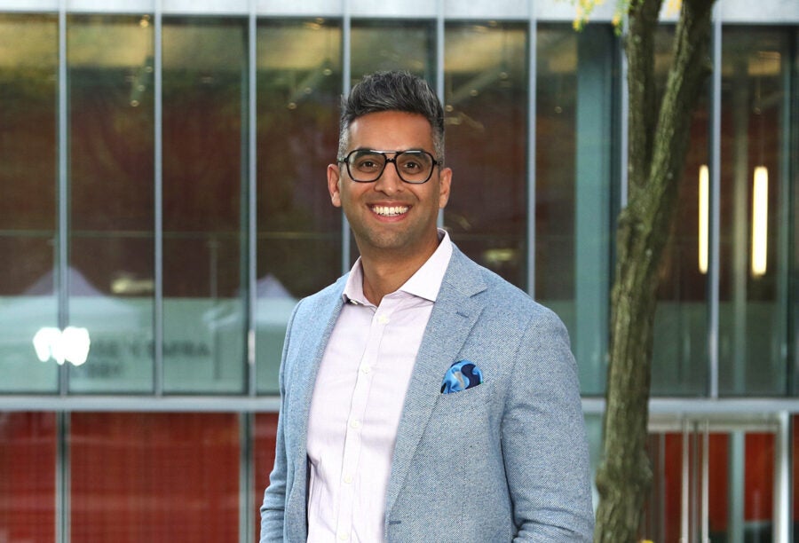 A person wearing glasses and a light gray blazer, standing outside in front of a modern glass building. Their expression is cheerful.