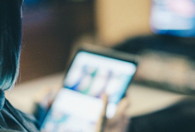 A person using a tablet, possibly streaming content, with a blurred TV screen in the background. The scene suggests leisure or relaxation.