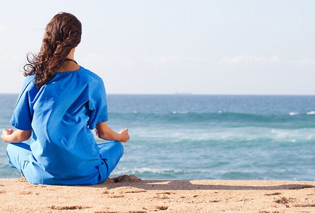 A person wearing blue scrubs is sitting cross-legged on a beach, facing the ocean, in a meditative pose.