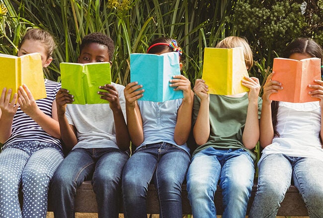 Five children sitting on a bench, each holding a colorful book in front of their faces, surrounded by greenery.