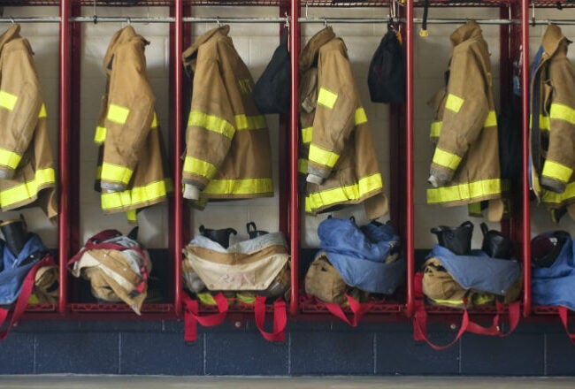 Firefighter uniforms, including jackets with reflective stripes, pants, and boots, organized neatly in red lockers.