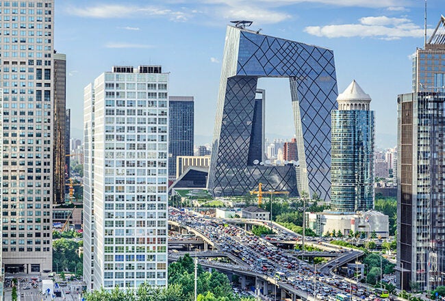 A cityscape featuring modern skyscrapers, with the distinctive, irregularly shaped CCTV Headquarters building in the center. A busy highway runs through the urban area.