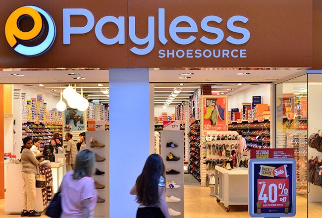 The image shows the storefront of a Payless ShoeSource store. It features a bright interior with a variety of shoes displayed, promotional signs, and people shopping. The store sign is prominently displayed above the entrance.