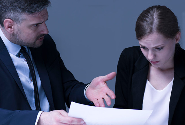A person in a suit aggressively gestures while showing a document to another person, who appears upset, in a dimly lit office setting.