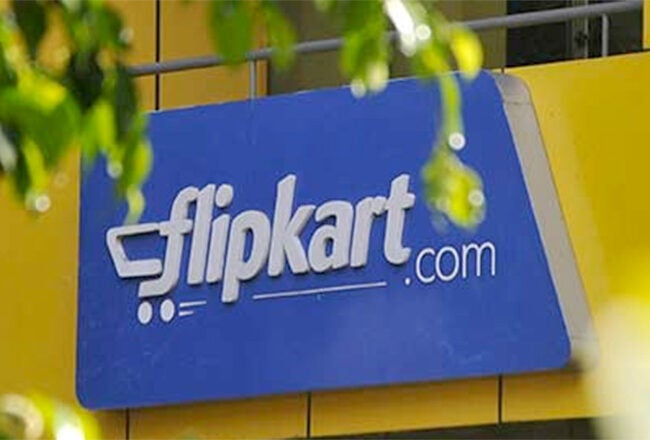 A blue sign with the Flipkart logo and a shopping cart icon, mounted on a yellow building facade, partially obscured by green leaves.