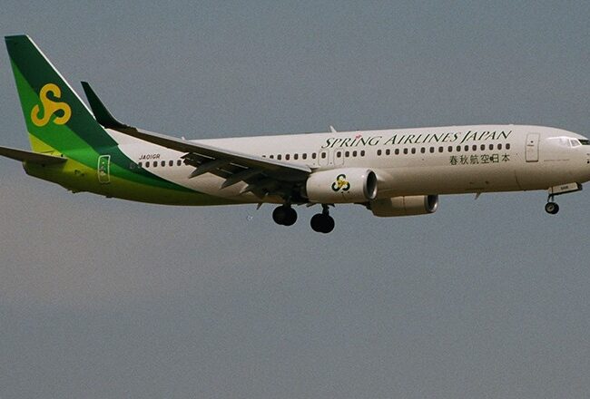 An airplane from Spring Airlines Japan in flight, with landing gear extended, displaying a green and yellow tail featuring the airline''s logo.