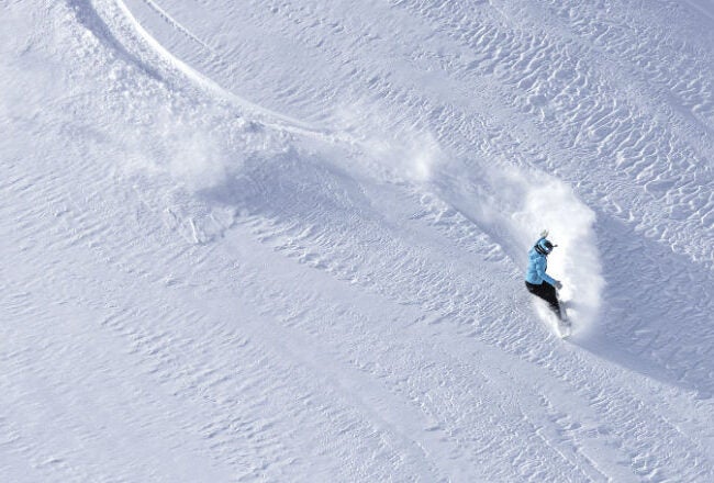 A snowboarder in a blue jacket gliding down a snowy slope, leaving a trail of powder behind.