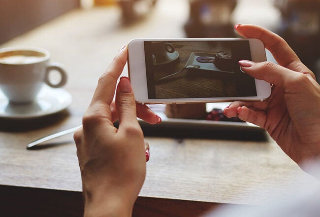 A person holding a smartphone, taking a photo of a cup of coffee on a wooden table. The scene captures a casual, cafe atmosphere.
