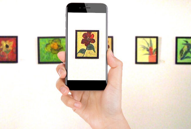 A person holds a smartphone taking a photo of a floral painting in an art gallery with several colorful flower artworks displayed on the wall.