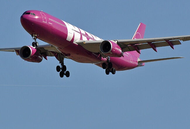A pink commercial airplane in flight against a clear blue sky, viewed from below with landing gear extended.