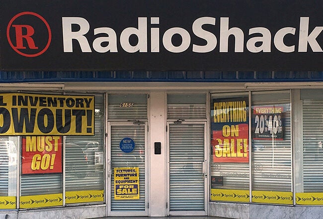 A RadioShack storefront with signs announcing a total inventory blowout sale. Large banners read "ON SALE" and "MUST GO!" indicating a possible store closing.