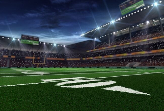 A football field under stadium lights, with a view of the end zone and yard lines. The stadium is filled with spectators, and the sky is dark.