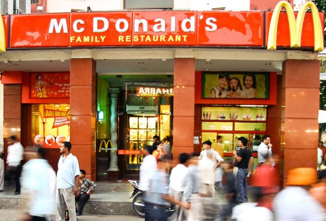 The image shows the exterior of a McDonald''s restaurant in a busy urban setting. Several people are walking past, creating a lively street scene.