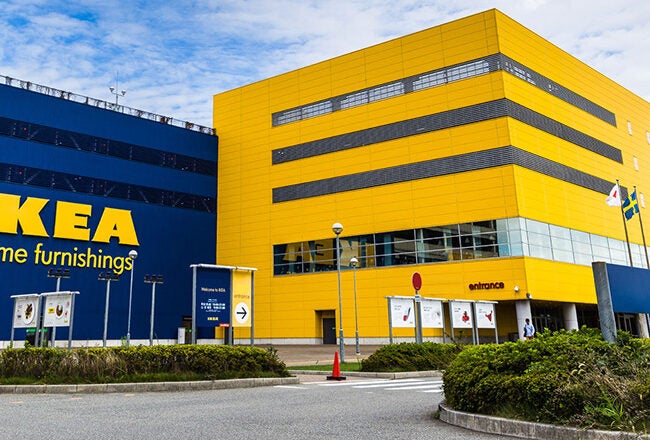 The image shows the exterior of an IKEA store with its distinctive blue and yellow color scheme. The building features the IKEA logo and signage indicating home furnishings and parking.