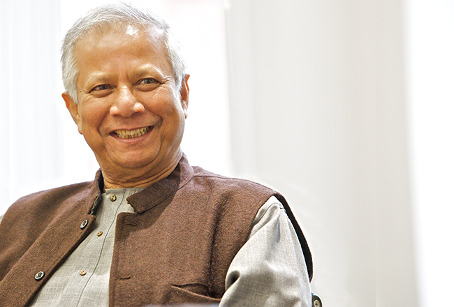 An individual smiling, wearing a gray shirt and brown vest, sitting indoors. The background is softly blurred, suggesting a warm and relaxed atmosphere.