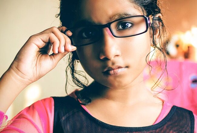 A young person wearing glasses and a colorful shirt, adjusting their glasses while looking directly at the camera. The background is softly focused with vibrant colors.