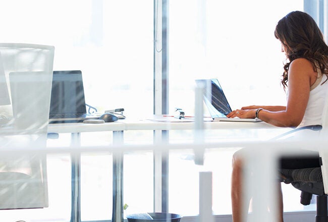 A person seated at a modern office desk, working on a laptop, in a bright office space with large windows.