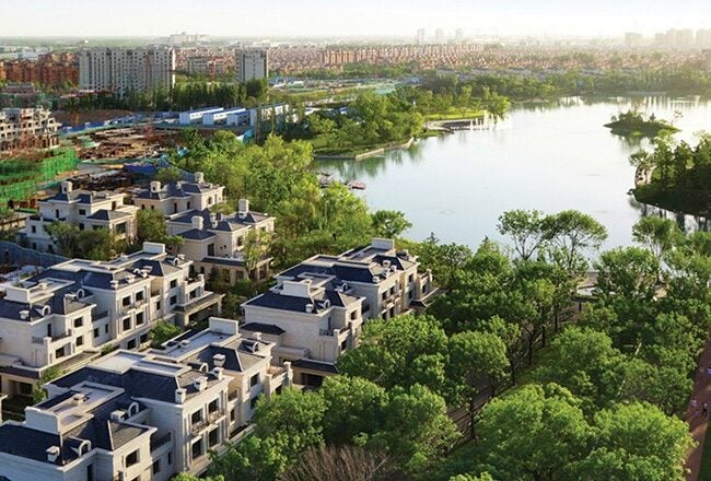 Aerial view of a residential neighborhood with modern houses, lush greenery, and a nearby lake. In the background, there are high-rise buildings.