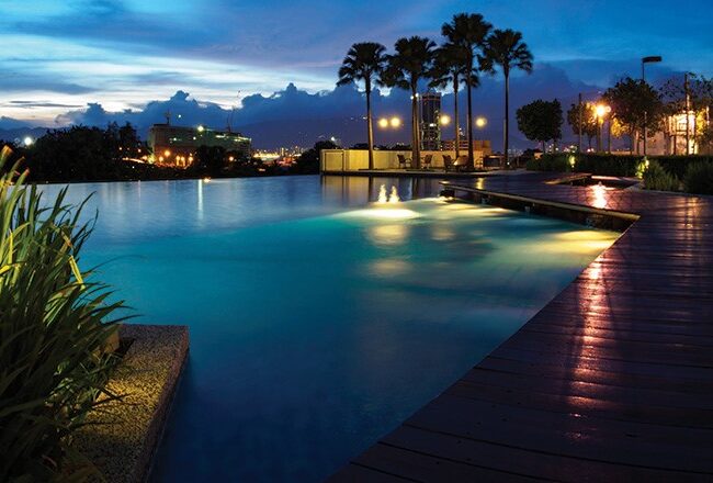 A beautiful night view of an illuminated infinity pool with surrounding palm trees and city lights in the background.