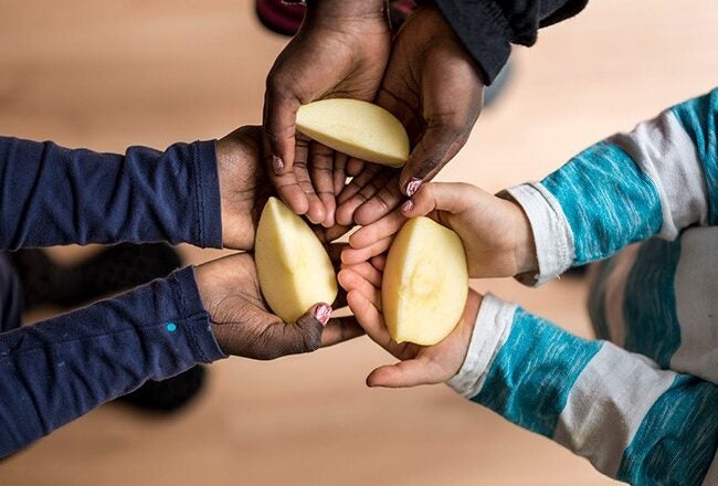 Three pairs of hands holding cut pieces of a yellow fruit, possibly a mango, symbolizing sharing or cooperation.