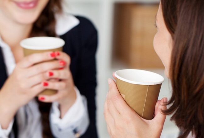 Two people holding coffee cups while sitting across from each other, possibly in a workplace setting, suggesting a casual conversation or break.