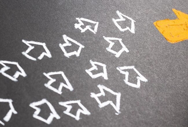 A hand drawing an arrow with chalk on a blackboard. Most arrows are white, pointing left, while one larger yellow arrow points right, symbolizing leadership or individuality.