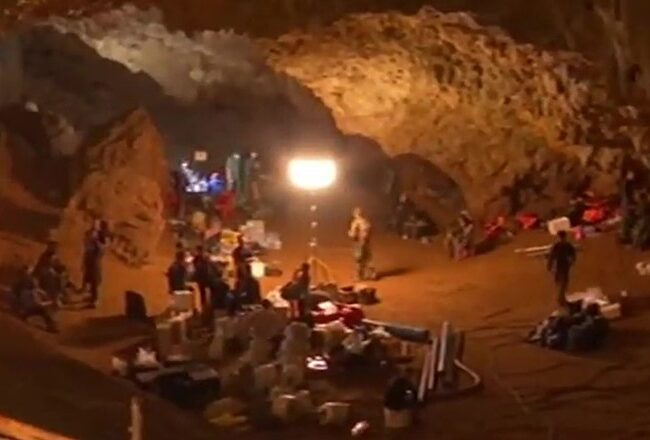 A large cave with people inside, illuminated by bright lights. Some are working, while others are sitting or lying down among scattered equipment and supplies.