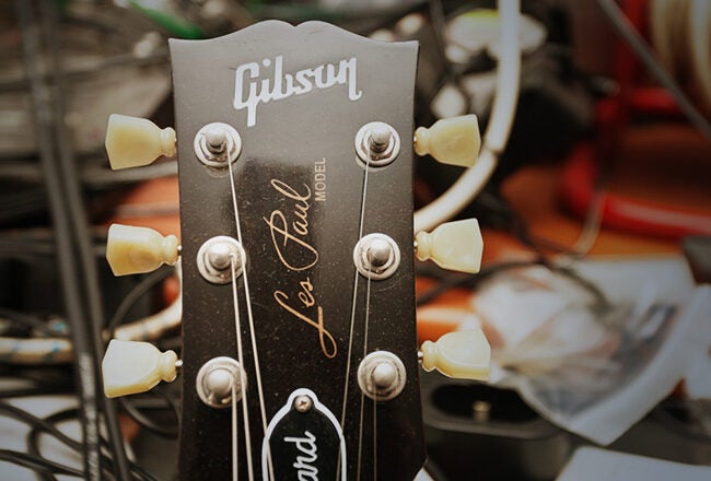 The image shows the headstock of a Gibson Les Paul guitar with tuning pegs, set against a background of audio cables and equipment.