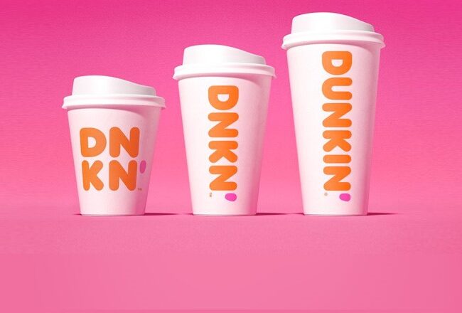 Three Dunkin'' coffee cups of varying sizes with pink and orange branding are on a pink background.