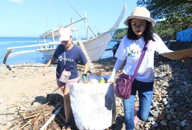 Two people cleaning up a rocky beach area, carrying a bag of collected trash, with a boat and ocean in the background.