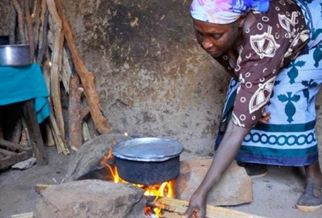 A person cooking over an open fire with a pot, inside a rustic setting. Wood is used as fuel, and there''s a table with kitchen items nearby.