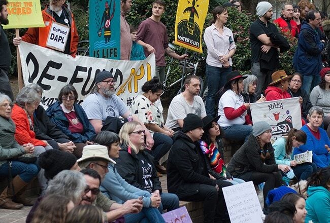 A group of people gathered for a protest, holding signs with messages such as "DeFund DAPL," "Water is Life," and "Respect the Treaties." They are seated and standing, some wearing colorful clothing and hats. The setting appears to