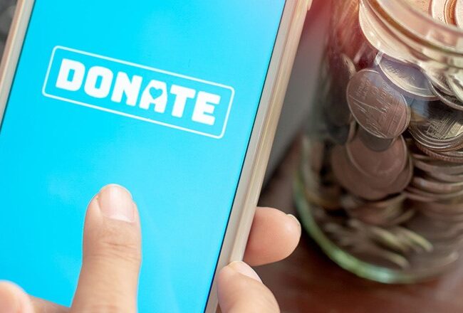 A person holding a smartphone with a "DONATE" button displayed on the screen next to a jar filled with coins.